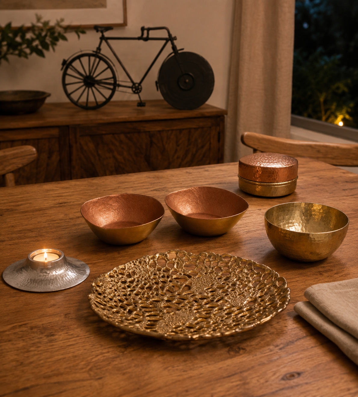 A set of decorative brass and copper bowls and a textured gold tray arranged on a wooden dining table, with a lit candle holder nearby in a warm, dimly lit home interior.